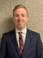 Headshot of a man smiling in a navy blue suit, white dress shirt, and maroon patterned tie.