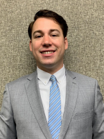 Headshot of a man smiling in a light gray suit, white dress shirt, and light blue striped tie.