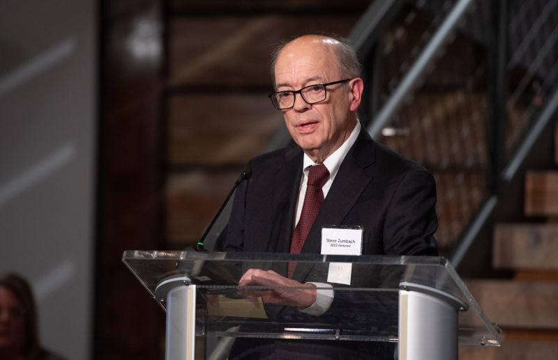 Man speaking at a podium in a black suite, white collar